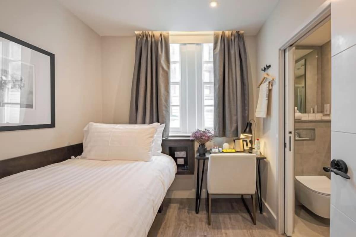 Una camera d'albergo con un letto, una scrivania con una sedia, un tavolino con una lampada e un bagno visibile attraverso una porta aperta.