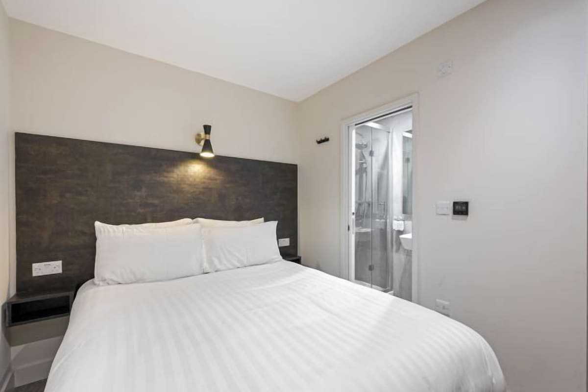 Una camera d'hotel moderna con un letto grande, una testiera scura, un comodino e un box doccia in vetro.