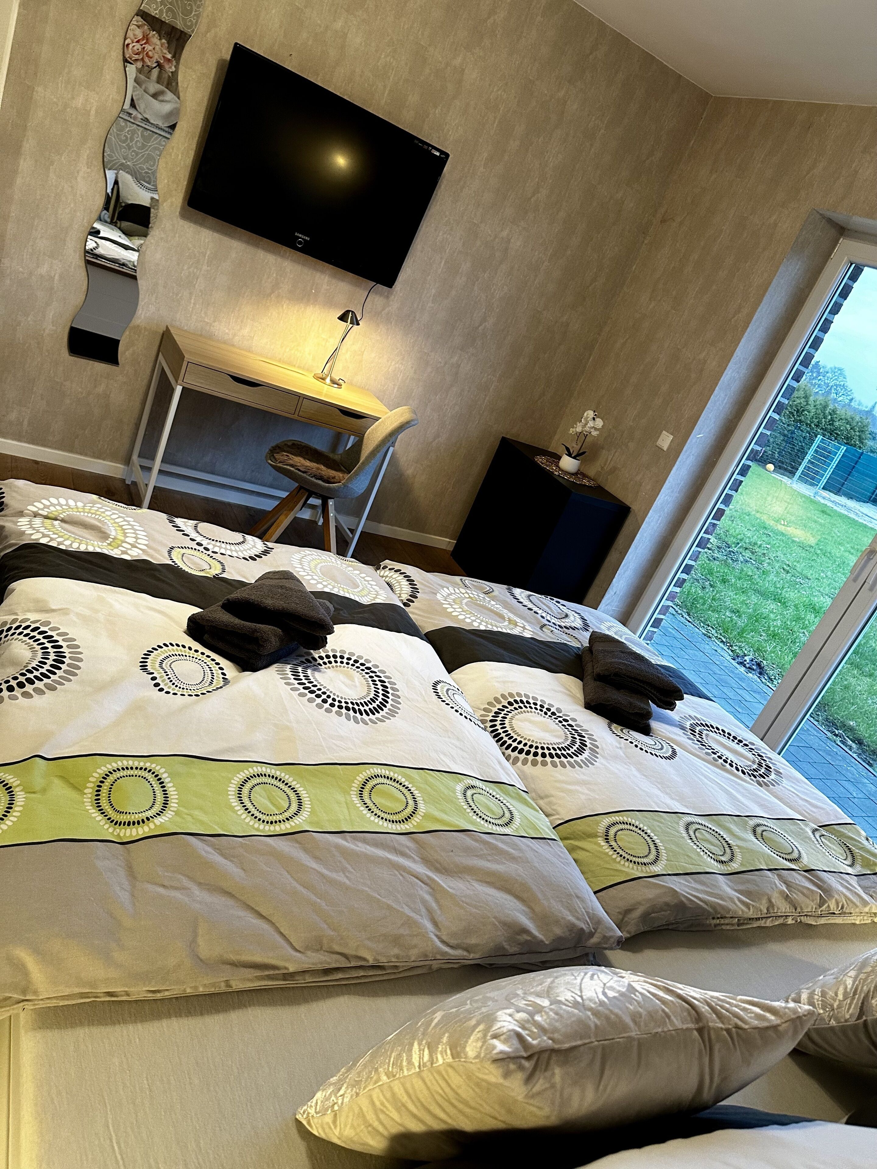 Ein Schlafzimmer mit einem großen Bett, einem Schreibtisch, einem Sessel, einem Fernseher und Blick ins Freie.
