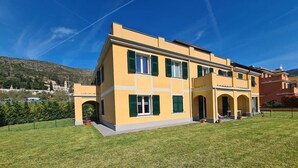 Un edificio de dos pisos con fachada amarilla y contraventanas verdes, que cuenta con una entrada central con arco y un pequeño porche.