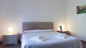 Una cama bien hecha con sábanas y toallas blancas, dos mesitas de noche con lámparas, y una pequeña planta en maceta sobre una de ellas.