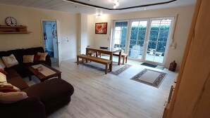 Ein Wohnzimmer mit einem dunkelbraunen Sofa, einem Holztisch und einem Esstisch mit Stühlen. Eine Tür führt in eine Küche, und ein großes Fenster bietet Blick auf viel Grün draußen.