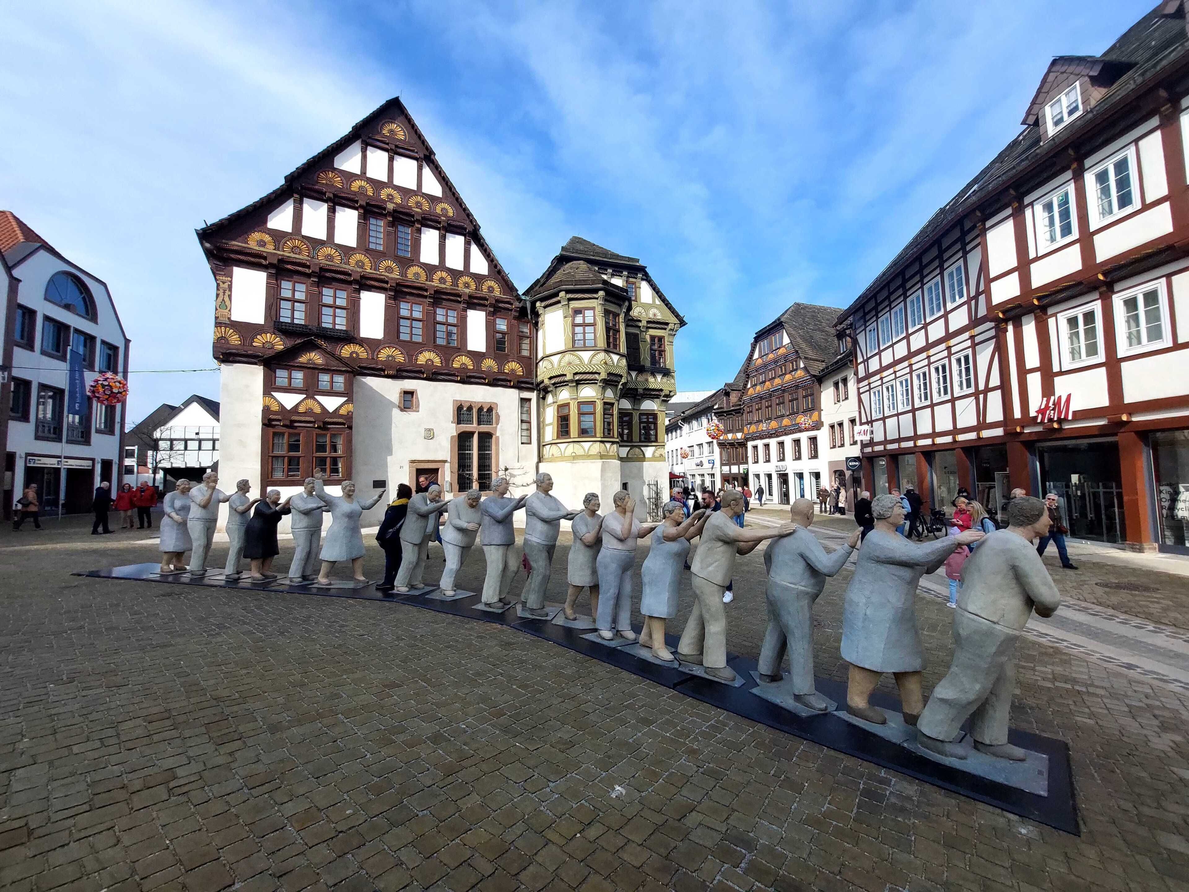 A cobblestone square with traditional half-timbered buildings, a statue of people in period clothing, and a clear blue sky.