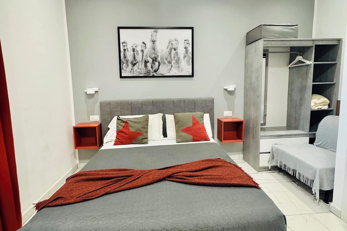A modern bedroom with a large bed, a grey headboard, a framed picture on the wall, a wardrobe, and a bedside table with a lamp.