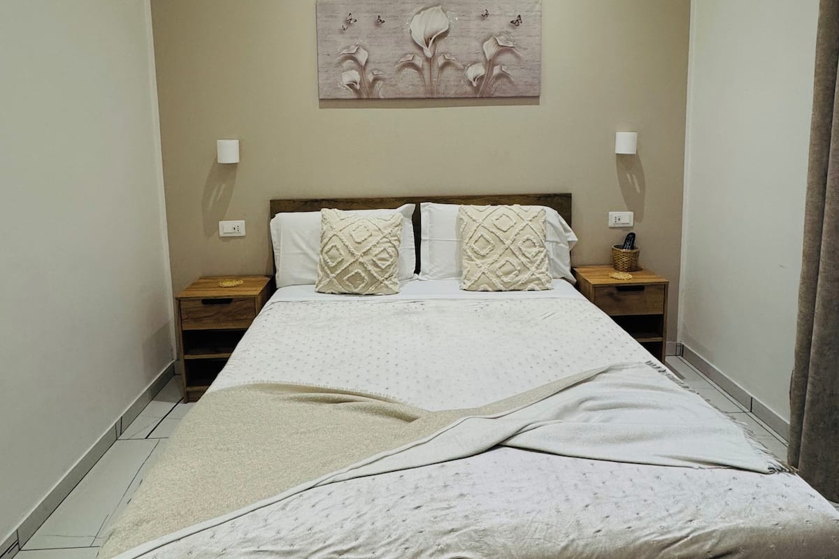 A bedroom with a bed, two nightstands, a wall-mounted lamp, and a painting.
