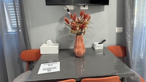 Un área de comedor con una mesa, sillas, un florero con flores, un televisor montado en la pared y una señal sobre la mesa.