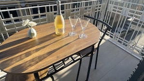 Una mesa de madera con una botella de vino y dos copas en un balcón con barandilla de metal.