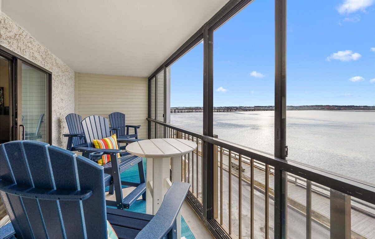 A balcony with two Adirondack chairs, a small table, and a view of a beach and pier.