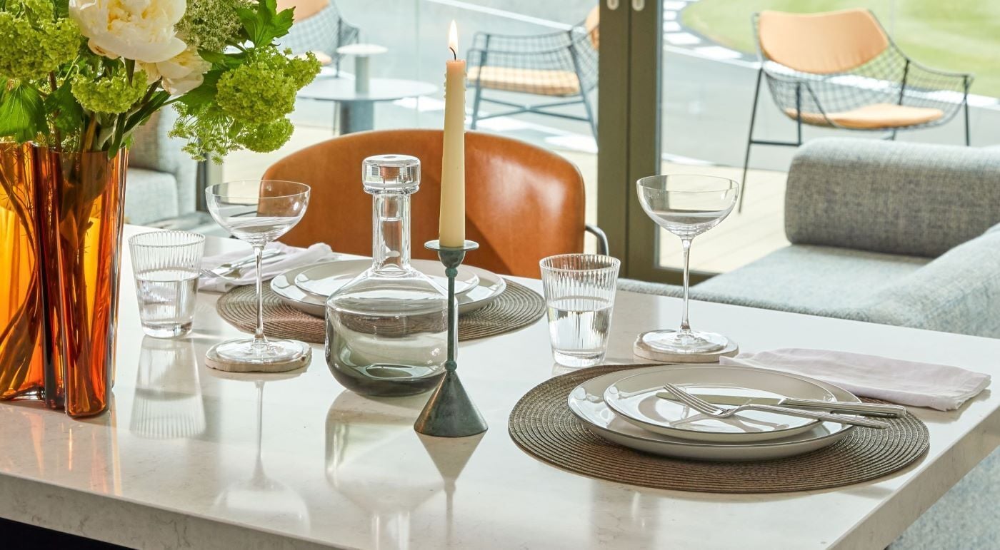 Una mesa de comedor dispuesta con platos, cubiertos, vasos, una vela y un florero con flores.
