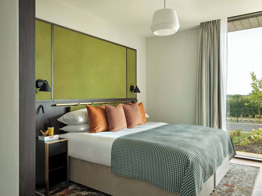 Habitación de hotel moderna con una cama grande, cabecera verde, dos lámparas de noche y vistas al exterior a través de un ventanal.