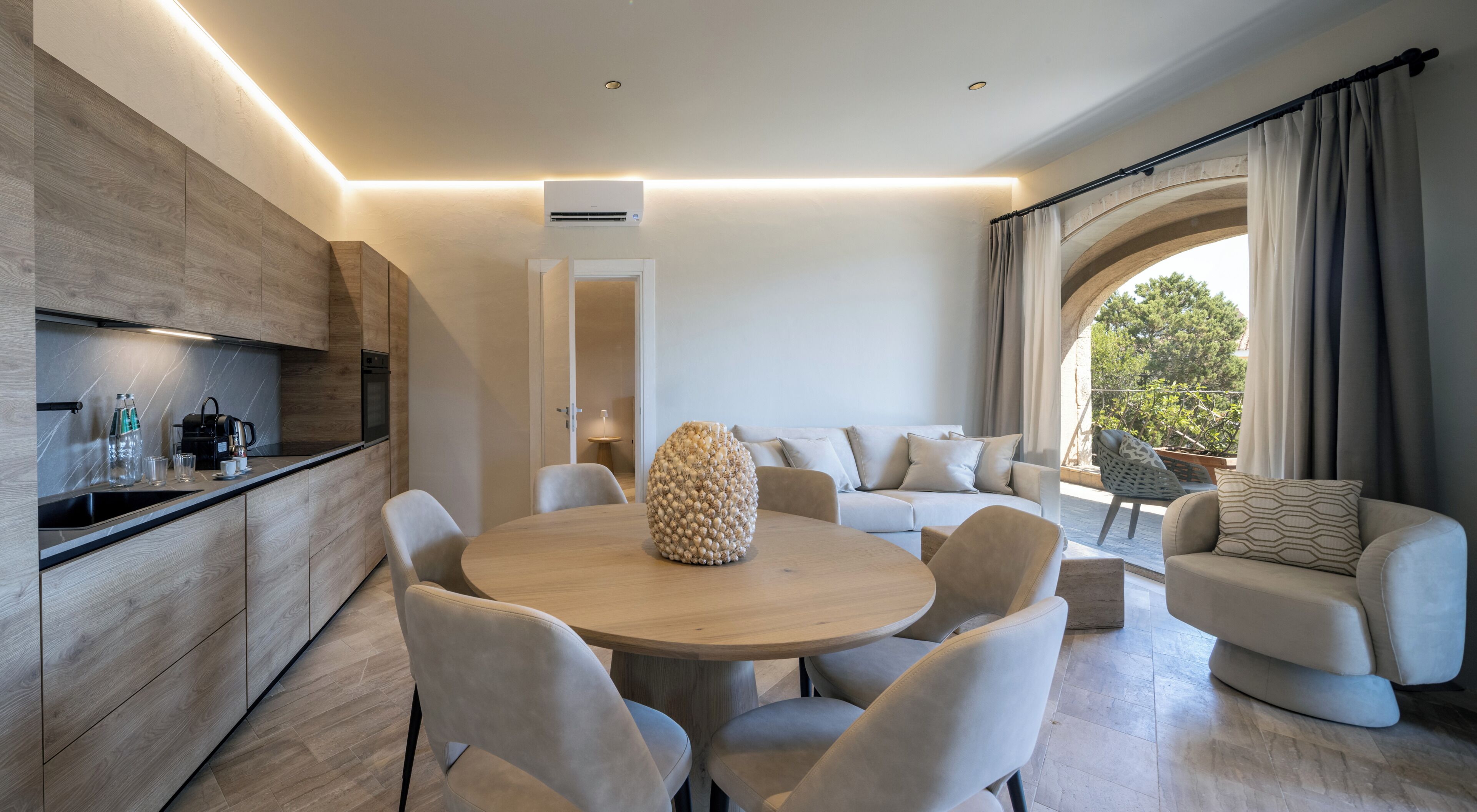 A modern living space with a round wooden table, beige sofas, and a kitchen area with a built-in oven.