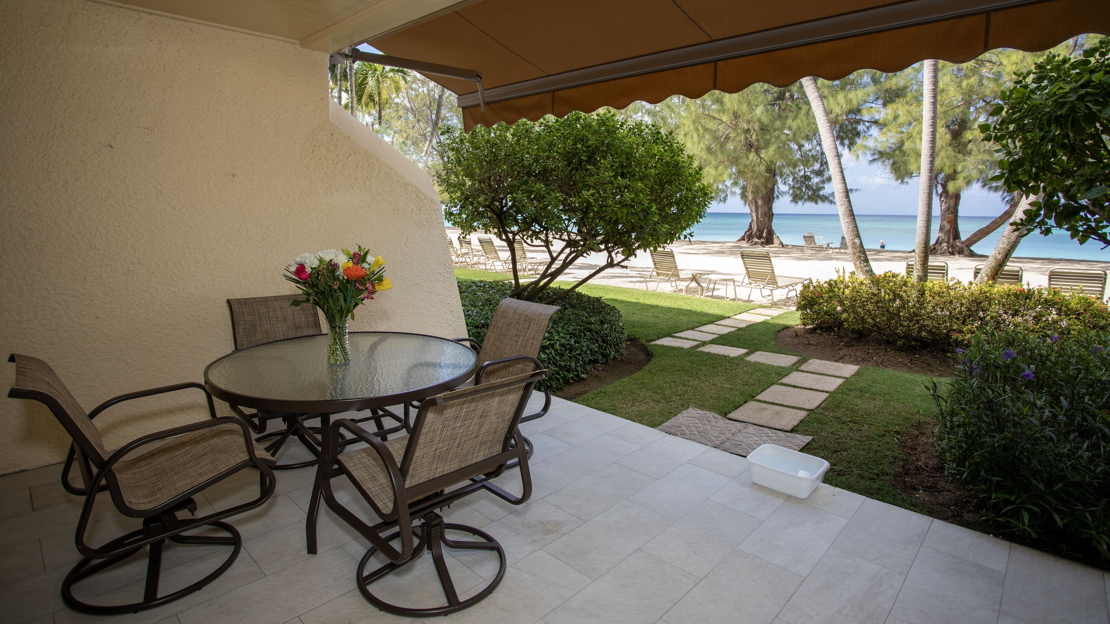 An outdoor patio with a round table and four chairs, a vase with flowers, and a view of the beach and palm trees.