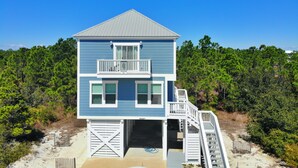 Ein Strandhaus mit zwei Etagen, einem Balkon, einer überdachten Veranda und einer Treppe, die zum oberen Stockwerk führt.