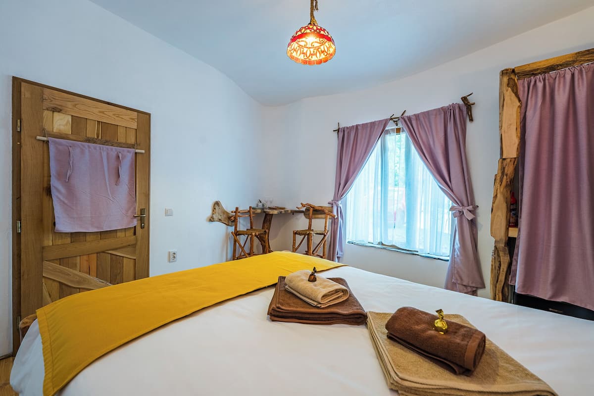 A bedroom with a bed, a wooden door, a window with purple curtains, and a hanging light fixture.