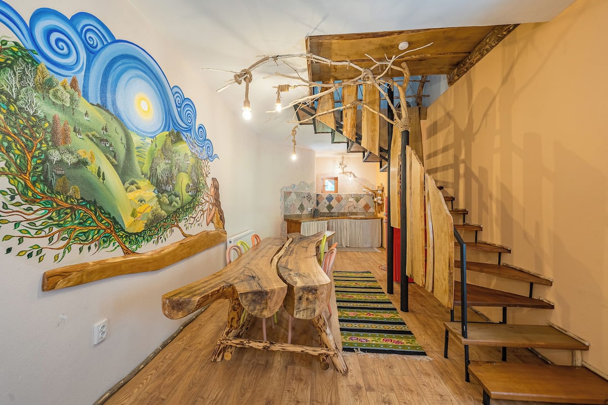 A dining area with a wooden table and chairs, a staircase, and a mural on the wall.