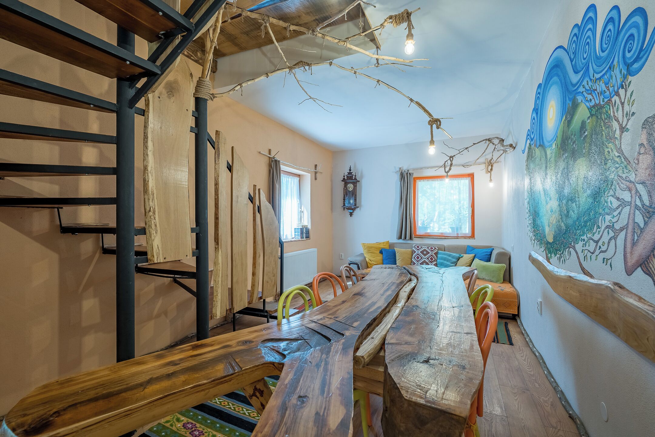 Un intérieur rustique avec une longue table en bois, des chaises colorées, un escalier et une fresque murale.