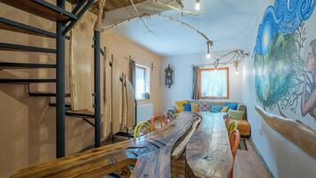 Un intérieur rustique avec une longue table en bois, des chaises colorées, un escalier et une fresque murale.