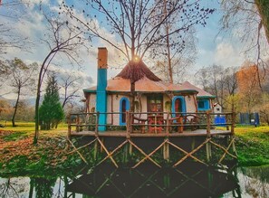A houseboat with a thatched roof, blue and white exterior, and a dock extending into a calm body of water. - Viewtopia - The Secret Land  (Poieni)