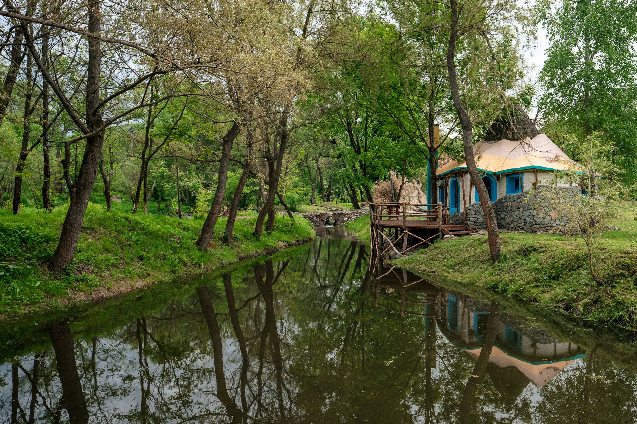 Un chalet rustique, doté de fondations en pierre et d’un toit bleu, entouré d’une végétation luxuriante et bordé par une rivière paisible.