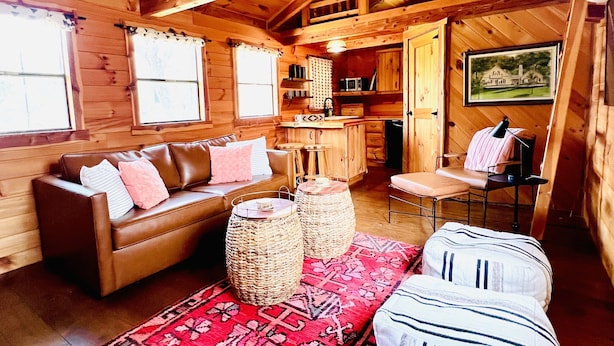 Acogedor interior de cabaña de madera con techo de madera, un sofá, otomanas, un tapete estampado y una zona de comedor.