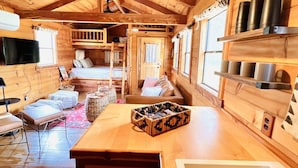 Acogedor interior de cabaña de madera, con comedor, cocina con estanterías y sala de estar con sofá y televisor.
