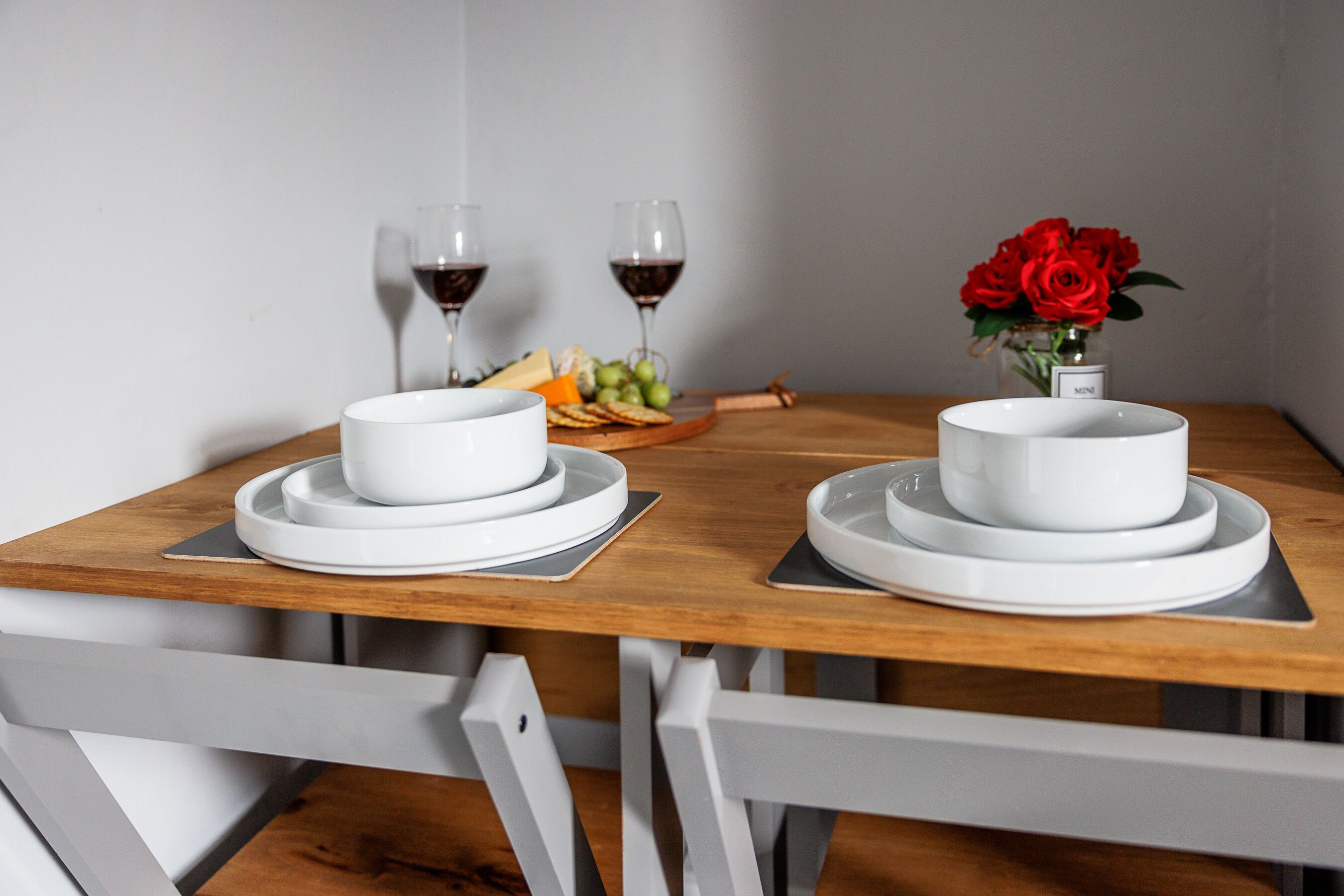 Une table à manger avec des assiettes et des bols blancs, deux verres de vin rouge, un bol de raisins, un bol de fromage et un vase rempli de roses rouges.