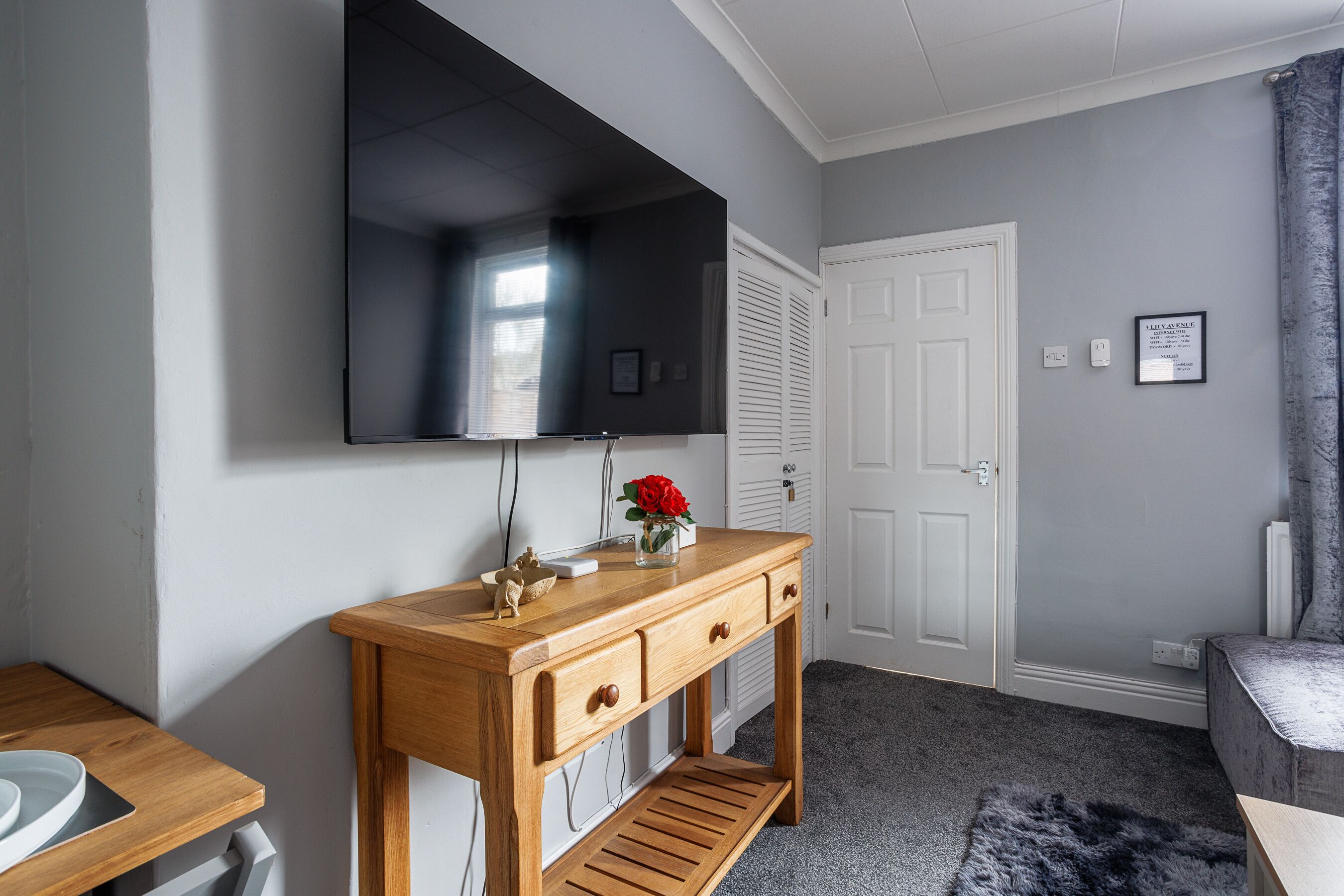 A room with a wooden console table, a flat-screen TV mounted above it, a white door, and a grey carpet.