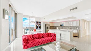 Amplia sala de estar con un sofá rojo, una mesa de centro de vidrio y una cocina moderna con armarios blancos y una isla iluminada con luz rosa.
