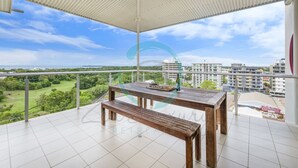 A balcony with a wooden table and bench, offering a view of a park and buildings. - Zen Ocean View Ph Spacious Living Pool Parking (Darwin)