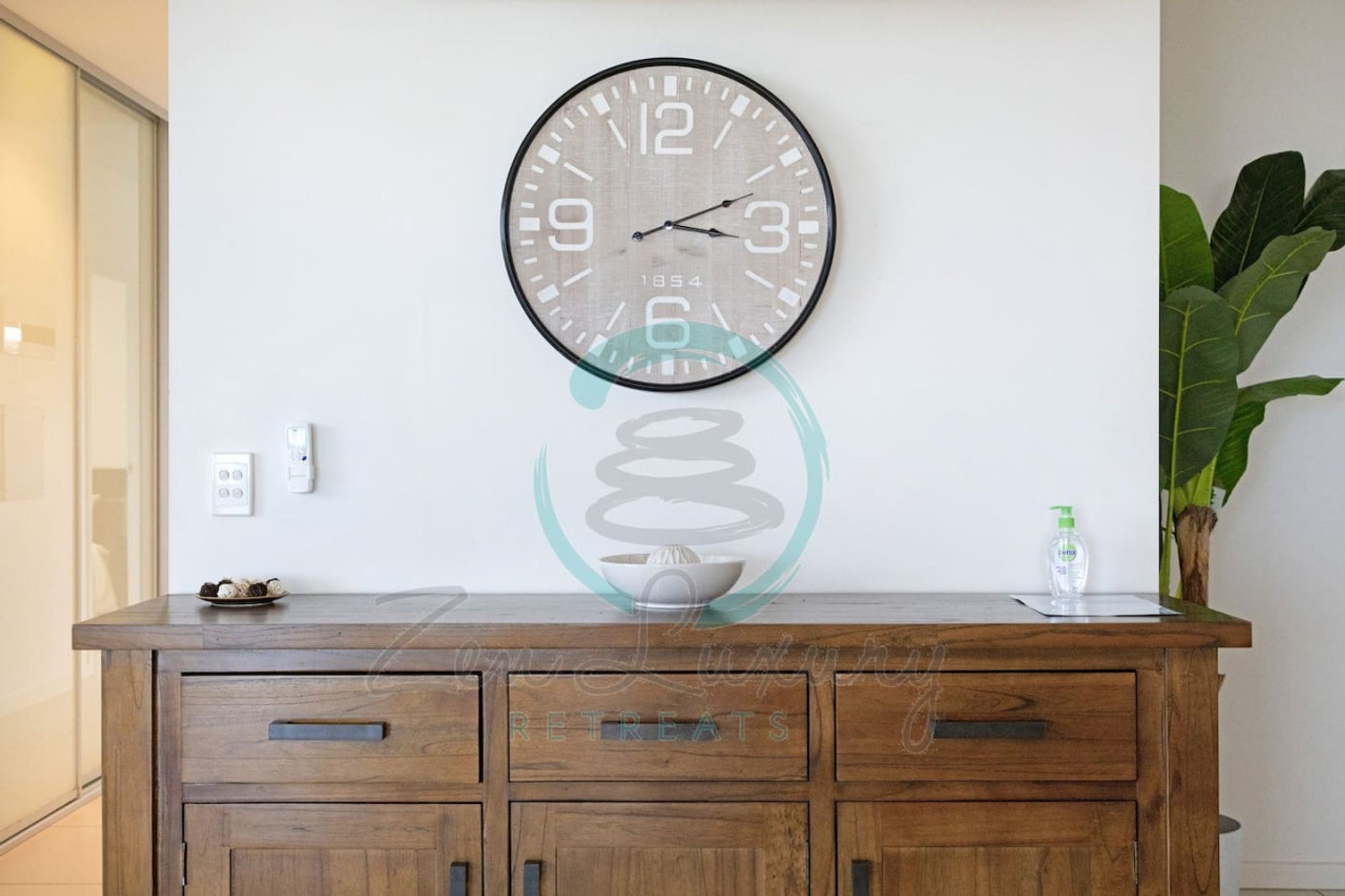 A wall clock with a minimalist design, a wooden cabinet with drawers, and a potted plant to the right.
