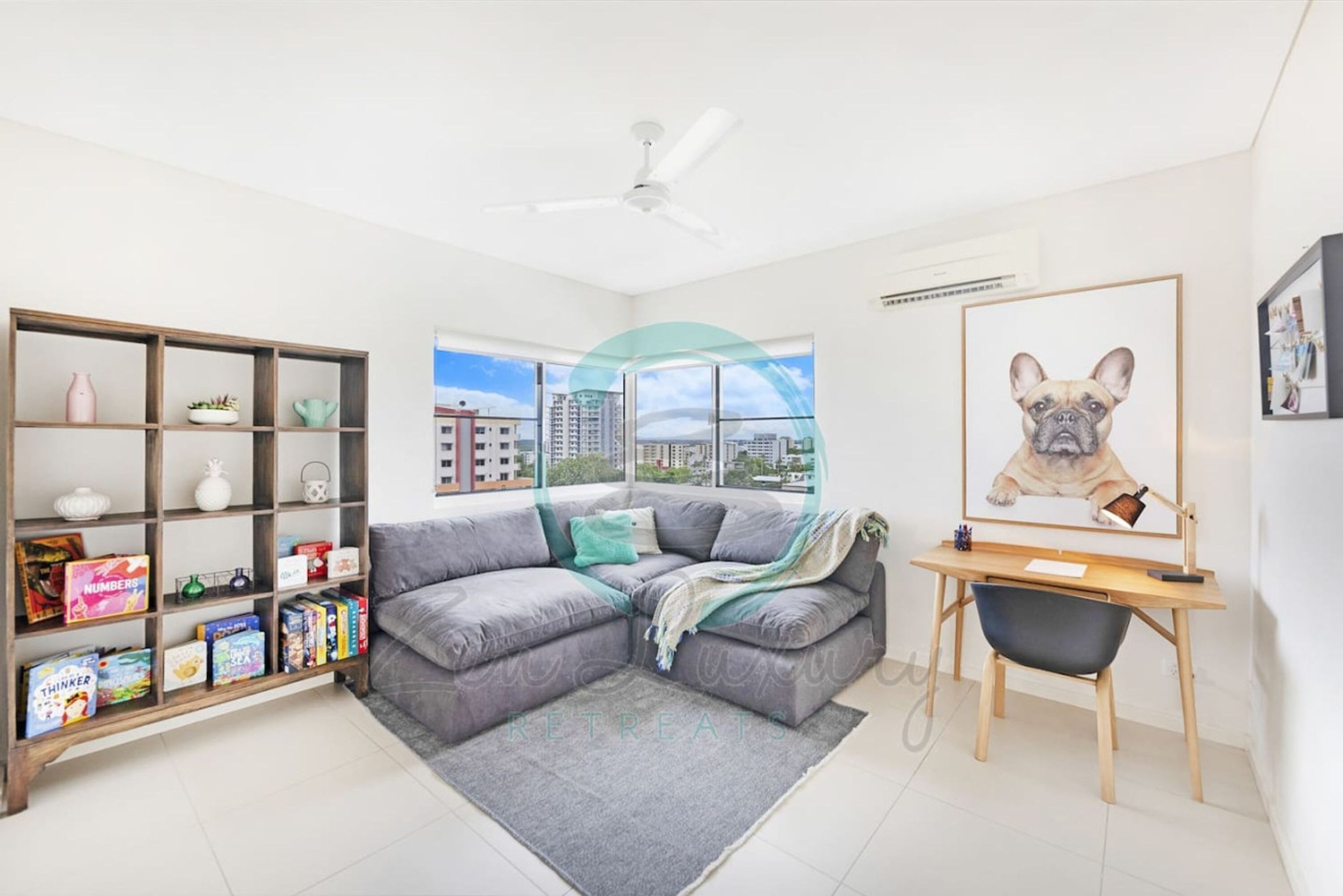 Uma sala de estar moderna com um sofá modular cinza, uma estante de madeira, uma escrivaninha com cadeira e uma janela grande com vista para a cidade.