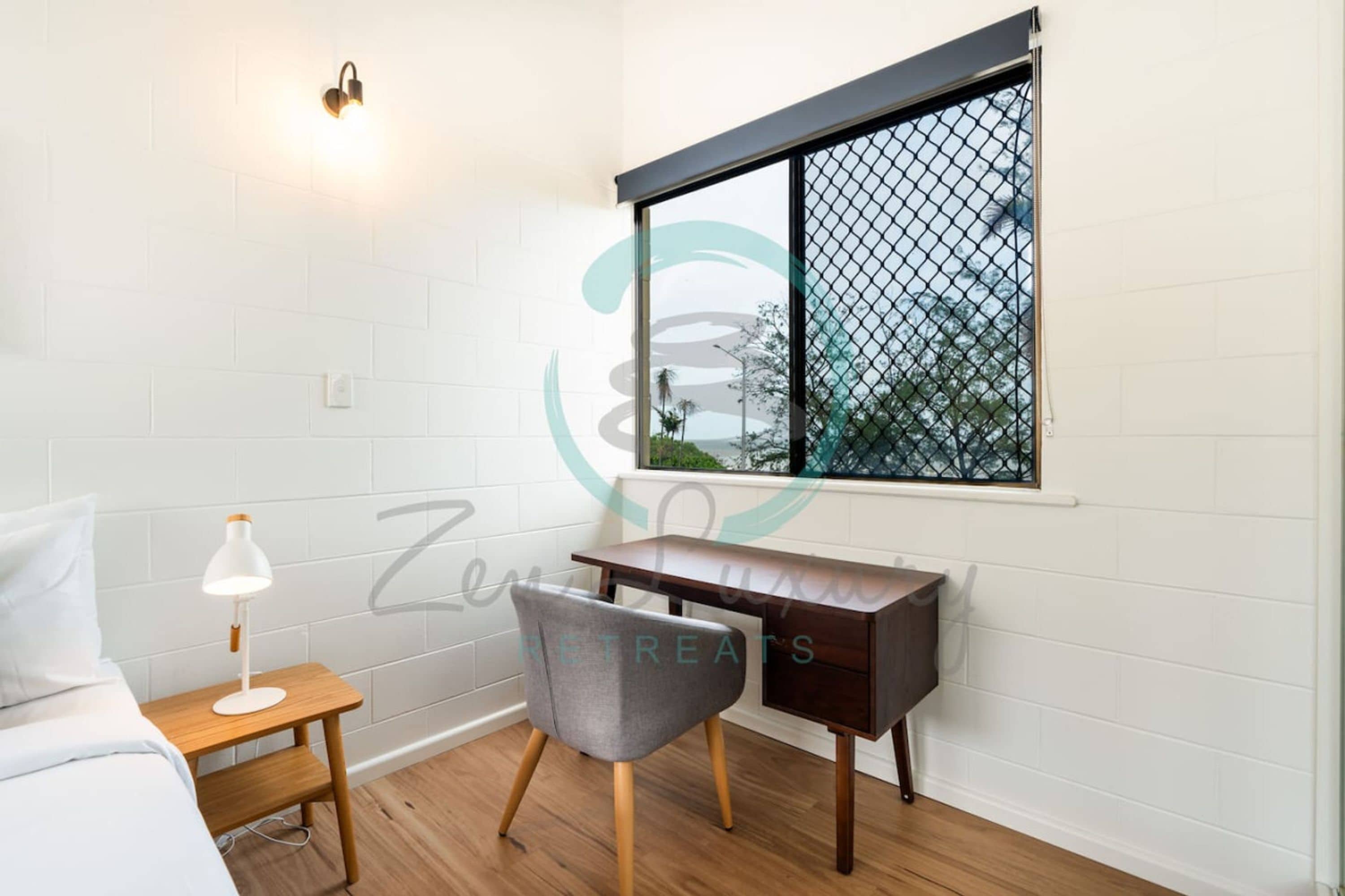 Un dormitorio con cama, una mesita de noche con lámpara, un escritorio y una silla. Hay una ventana que ofrece vistas a árboles y un edificio.