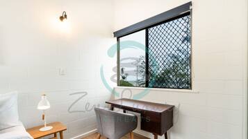 Un dormitorio con cama, una mesita de noche con lámpara, un escritorio y una silla. Hay una ventana que ofrece vistas a árboles y un edificio.