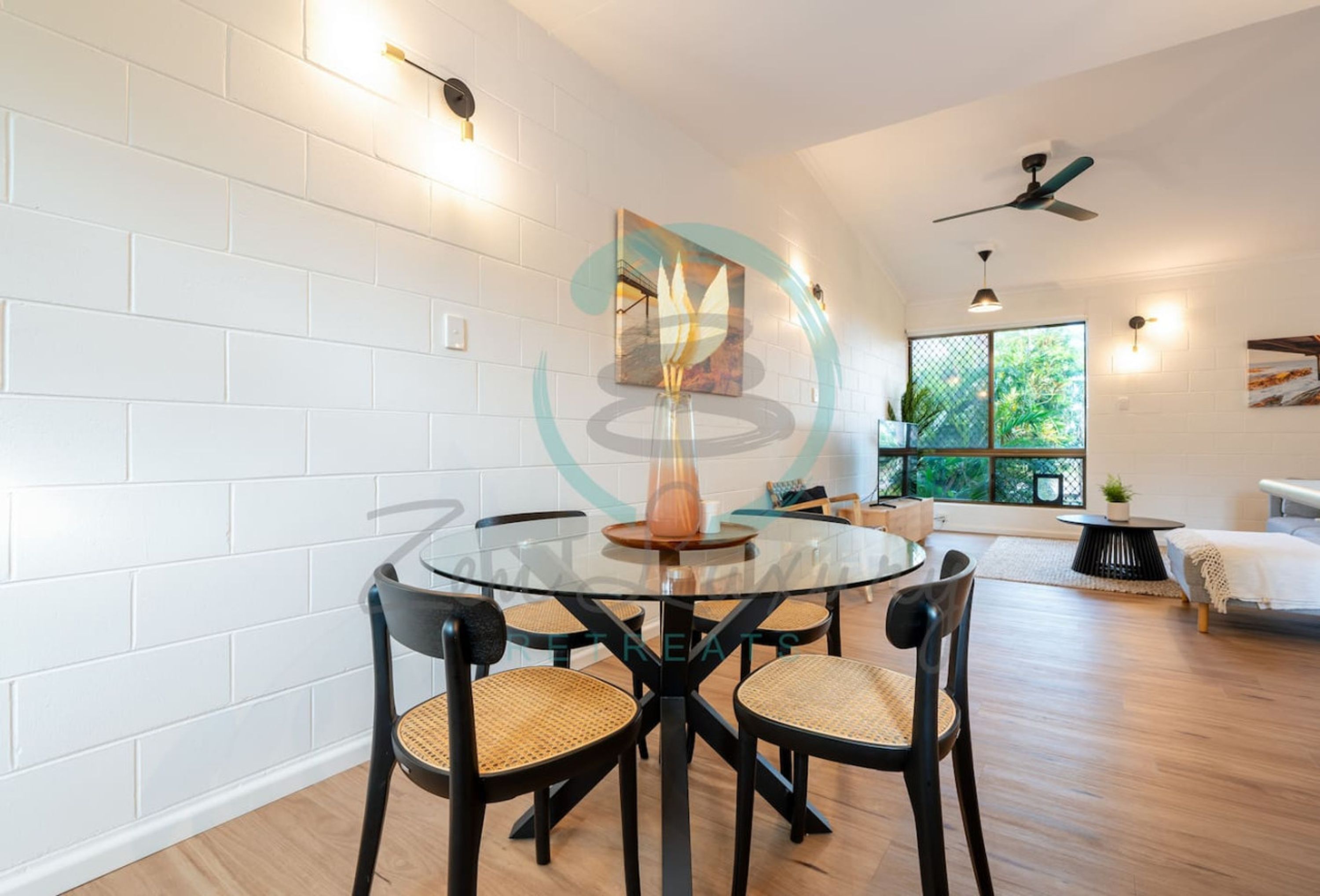 A modern dining area with a glass table and black chairs, white brick walls, and a ceiling fan.
