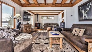 A cozy living room with a fireplace, wooden beams, and a large window overlooking a snowy landscape. - Stepaway Vacation Rentals- Lost at Last (St.Germain)