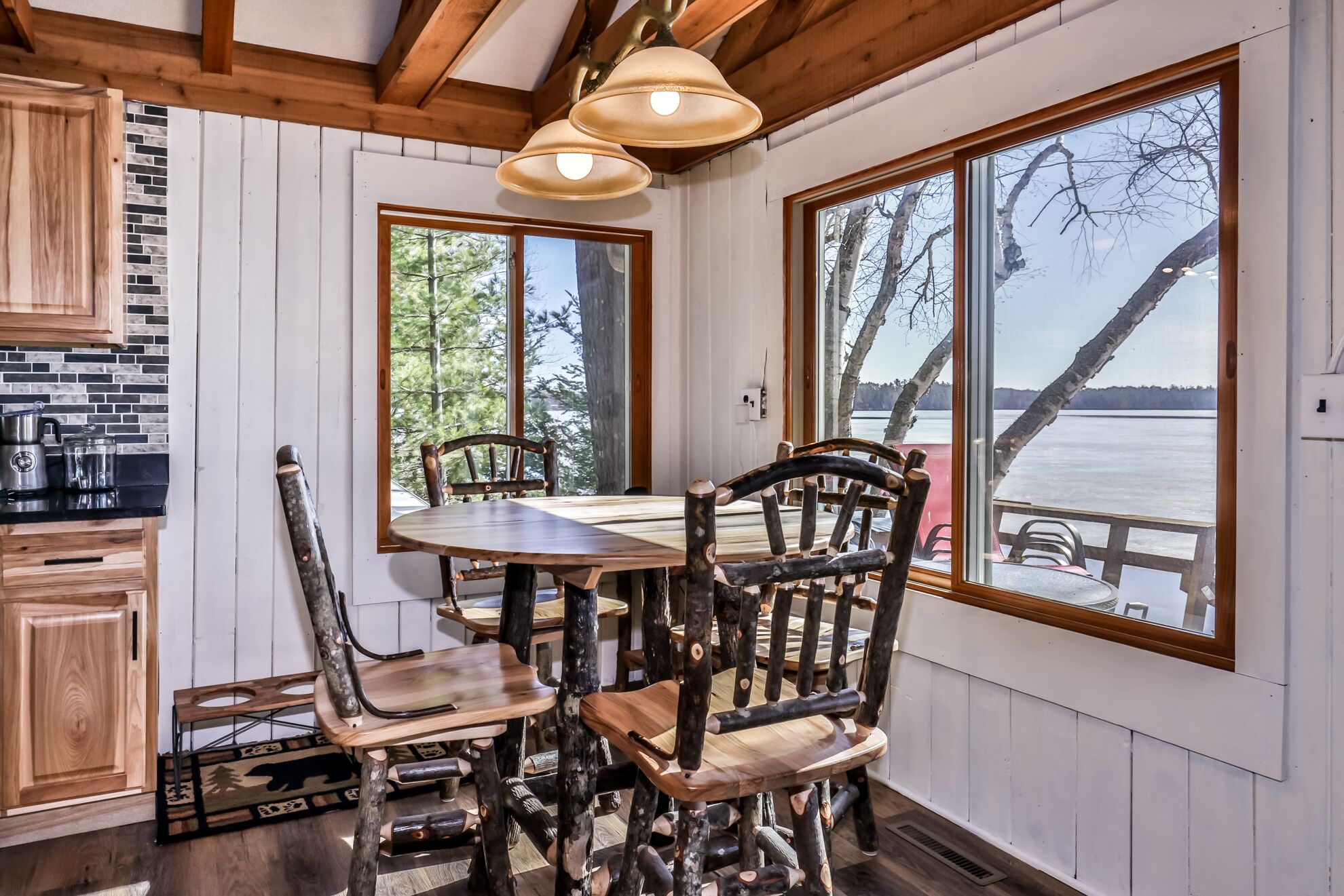 L’intérieur d’un chalet rustique est doté d’une table et de chaises en bois, de grandes fenêtres offrant une vue sur un lac, et d’un coin cuisine avec un mur de fond en briques.