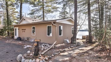 A small cabin with a metal roof, a wooden door, and a window. There is a pile of rocks in front of the cabin and a tree nearby.