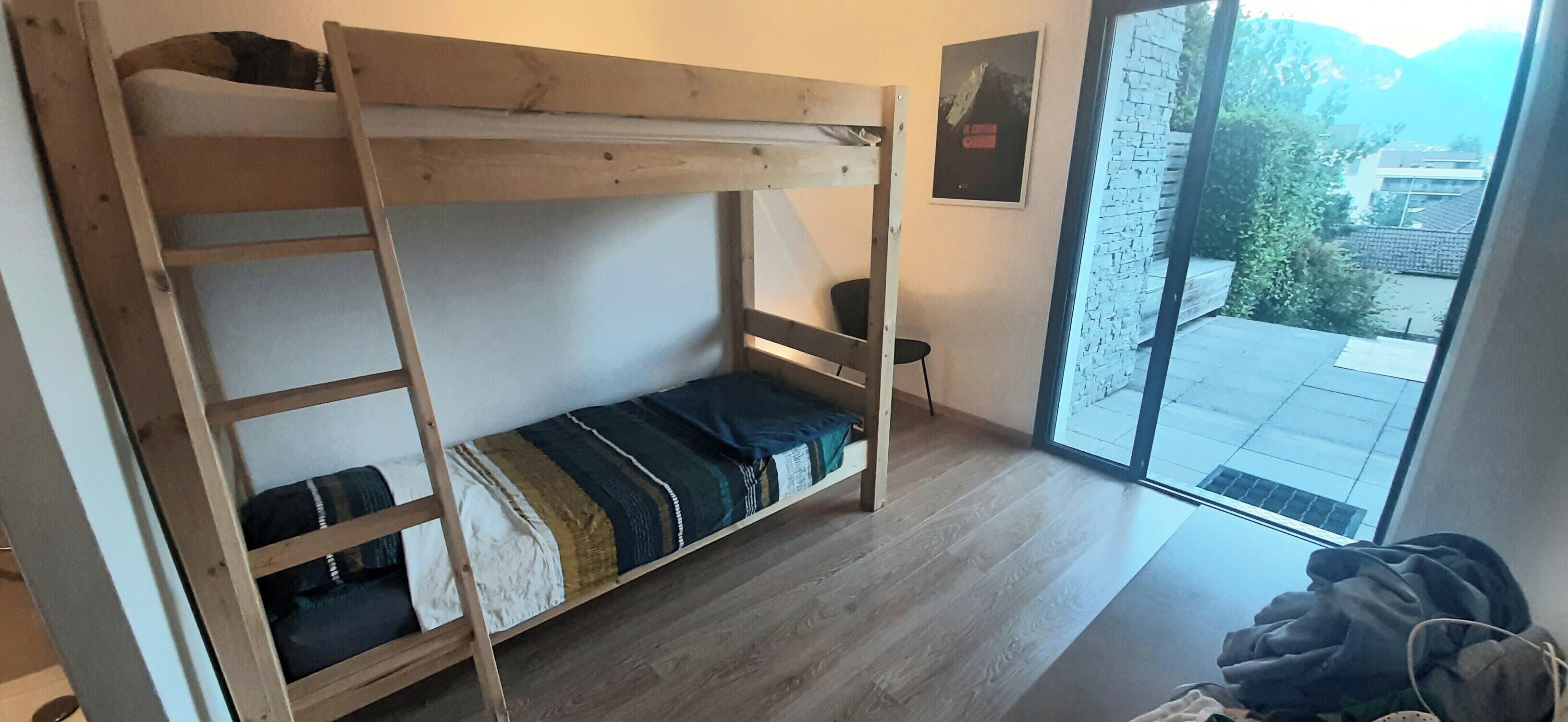 Un lit superposé avec une structure en bois et un matelas simple. Il y a un petit tabouret à côté du lit et une fenêtre donnant sur un immeuble et des arbres.
