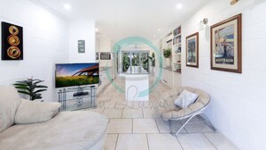 A modern living room with a flat-screen TV, a sofa, a coffee table, and a decorative archway leading to a dining area. - Zen Treetops Private Pool Outdoor Dining (Larrakeyah)