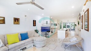 A modern living room with a sofa, a coffee table, and a TV. There is a ceiling fan, a kitchen area in the background, and a dining area with a table and chairs. - Zen Treetops Private Pool Outdoor Dining (Larrakeyah)