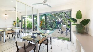 A modern dining area with a glass table and chairs, a ceiling fan, and a view of the outdoor garden. - Zen Treetops Private Pool Outdoor Dining (Larrakeyah)