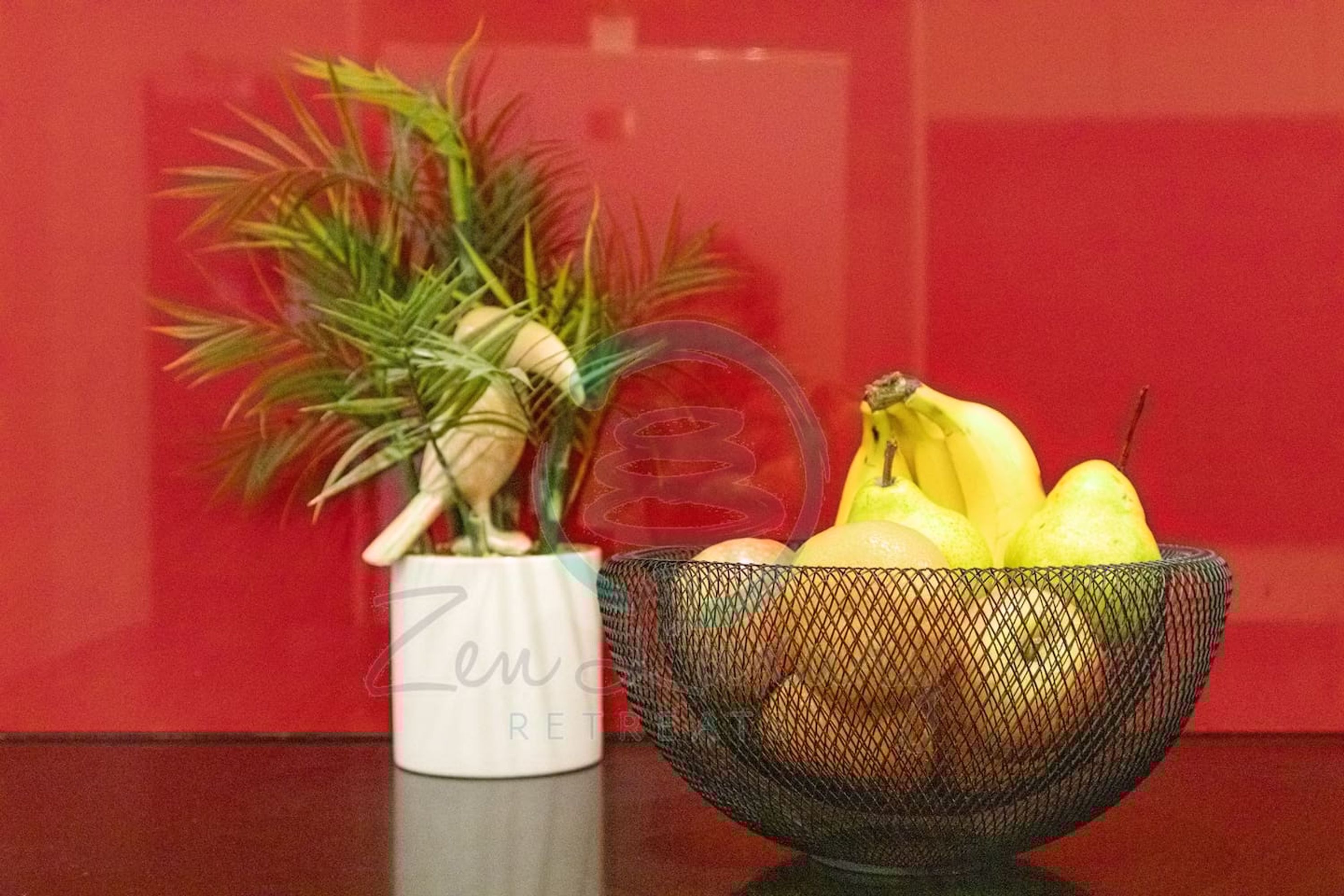 A white vase with a plant and a black mesh bowl containing fruits.
