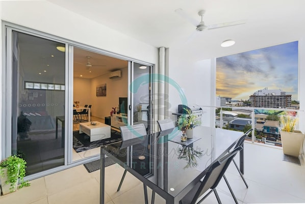 A modern dining area with a glass table and chairs, a ceiling fan, and a view of the cityscape through large windows.