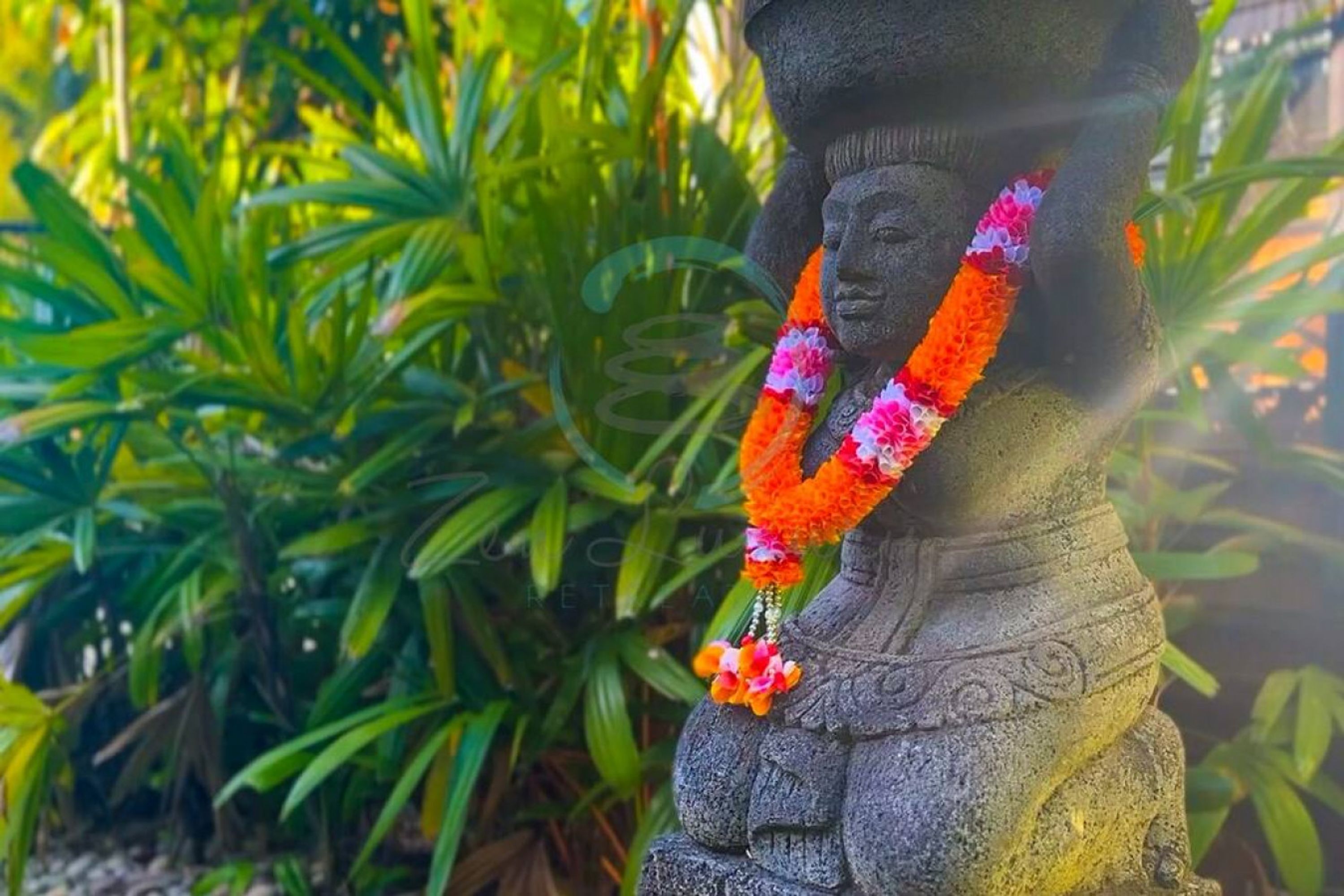 A stone statue with a floral garland, set against a backdrop of green foliage.