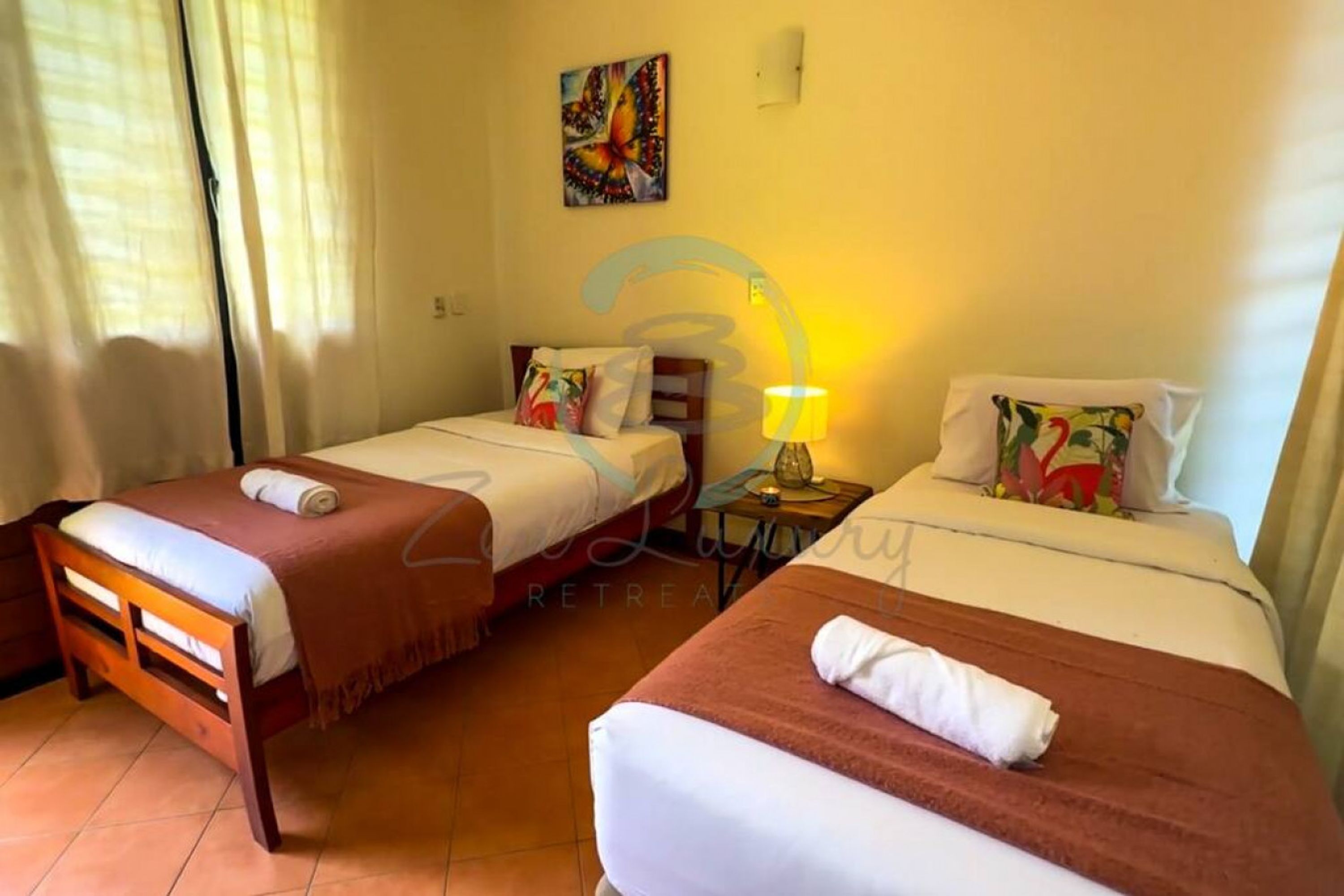 A hotel room with two single beds, a small table with a lamp, and a painting on the wall.