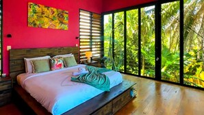 Ein Schlafzimmer mit einem großen Bett, einem Holz-Kopfende und Blick auf üppiges Grün durch große Fenster.