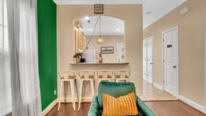 Un salon moderne avec un canapé vert, un coin bar avec des tabourets et une cuisine visible à travers une arche.