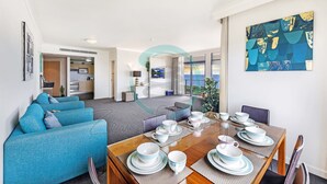 A modern living room with a dining area, featuring a sofa set, a dining table set with plates and cups, and a large abstract painting on the wall. - Rustic Sunset Horizon Oceanview Balcony Dining (Darwin)