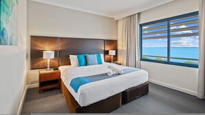 A hotel room with a large bed, two bedside tables, a headboard, a window with a view of the sea, and a painting on the wall. - Rustic Sunset Horizon Oceanview Balcony Dining (Darwin)