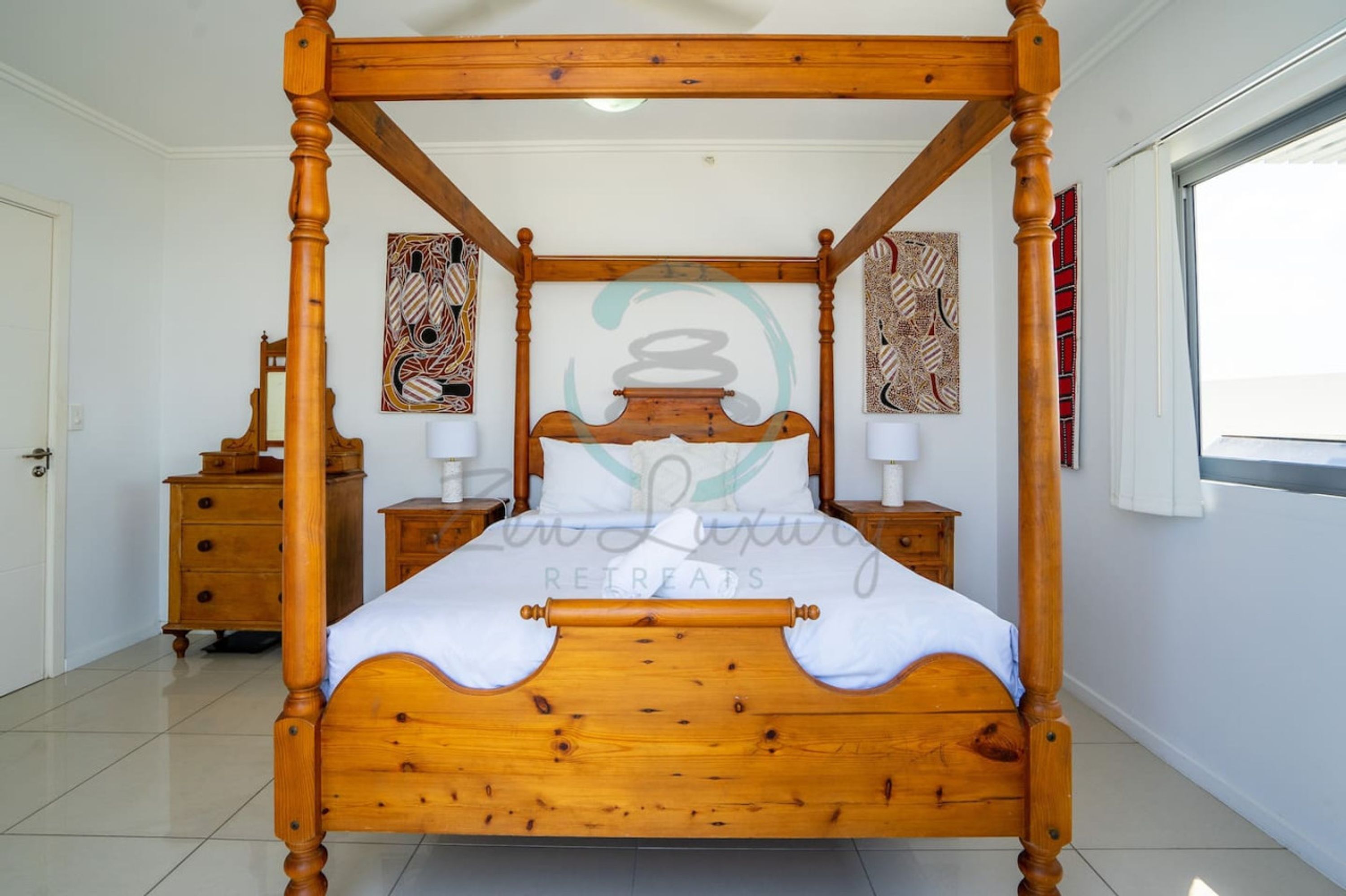 Una cama con dosel de madera y ropa de cama blanca en una habitación con paredes blancas y un ventanal grande.